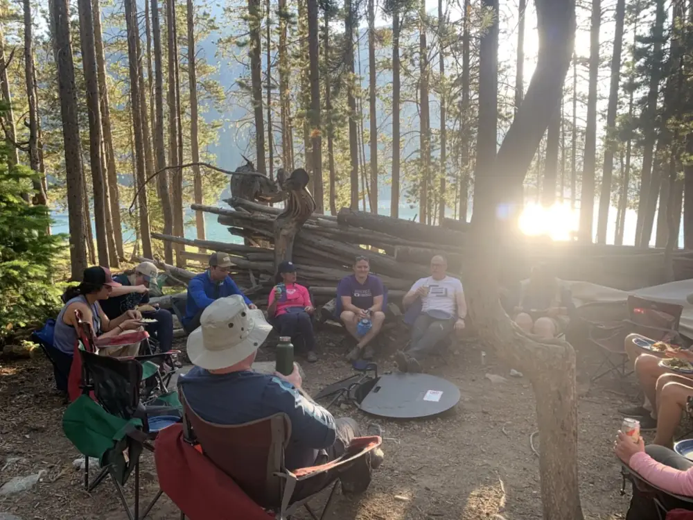 Group of approximately 6-7 people seated in camping chairs and campfire or cooking setup in center in Grand Teton National Park