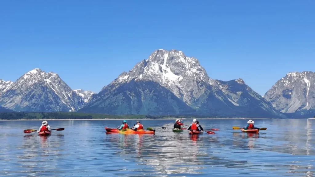 Six people in kayaks on calm lake water and red and orange kayaks with black paddles in Grand Teton National Park.