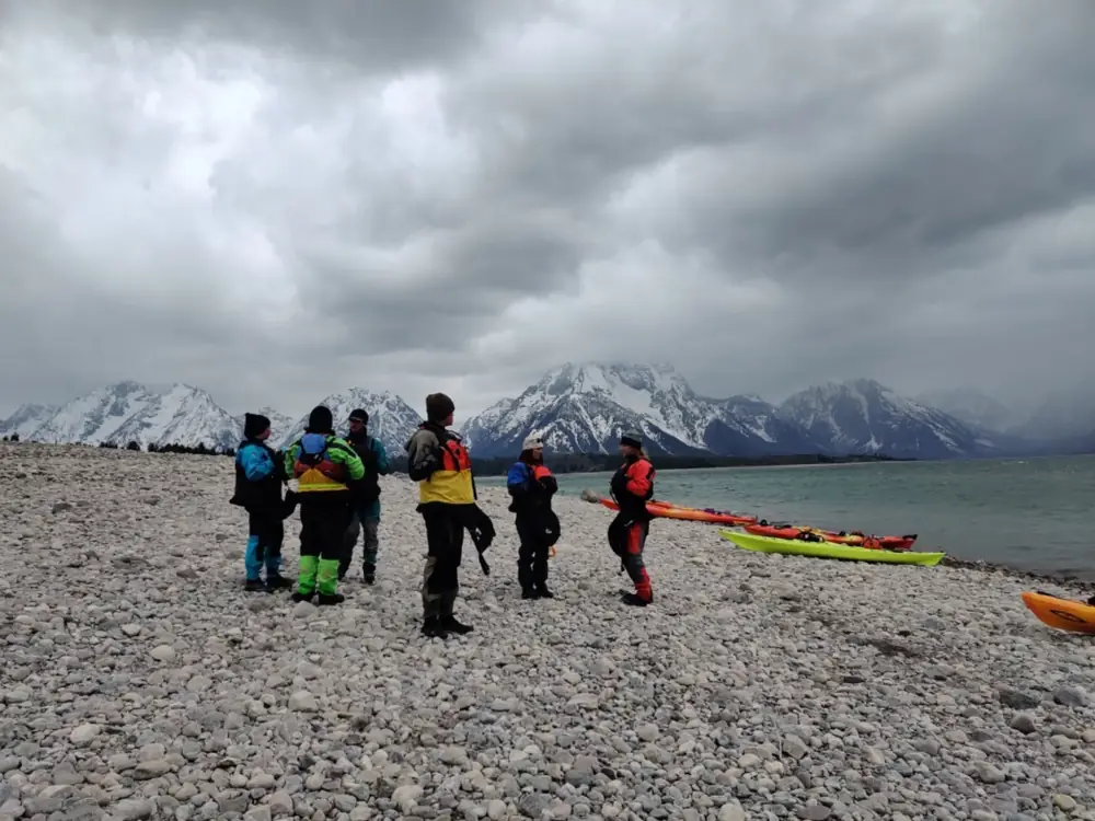Group standing together on beach, backs mostly to camera, wearing paddling gear in Snake River.