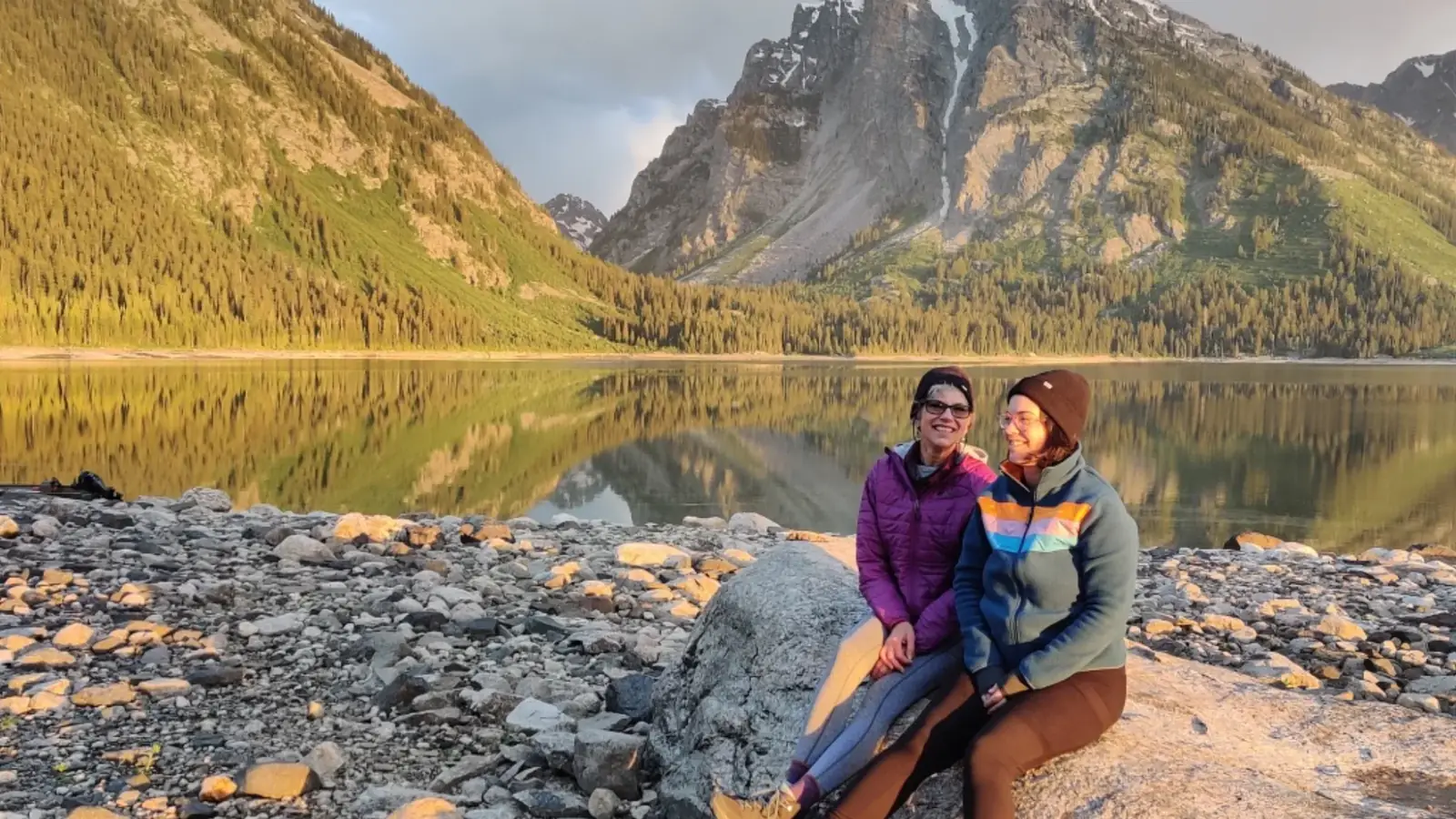 Two people seated on rocky shoreline and calm lake with mirror-like reflection in Grand Teton National Park.