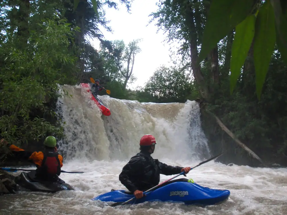 Three people in kayaks and one red kayak at the top edge of a waterfall in Jackson Hole.