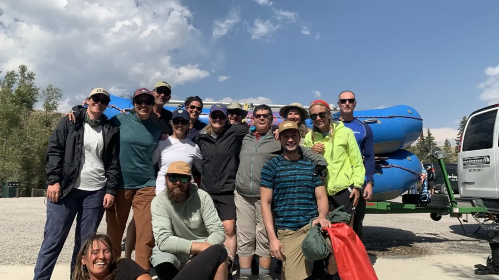 Group of approximately 16 people posing in parking lot and blue inflatable rafts on vehicle rack in background in Jackson Hole, Wyoming