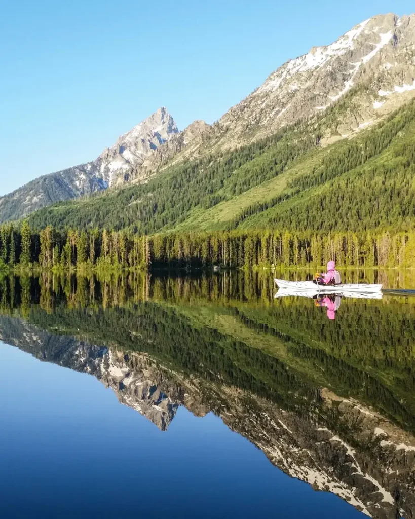 Calm lake with mirror-like reflection and mountain range with rocky peaks and patches of snow in Jackson Hole.