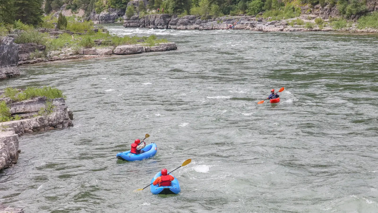 Three people in inflatable kayaks on clear river water and two blue inflatable kayaks in foreground in Jackson Hole.
