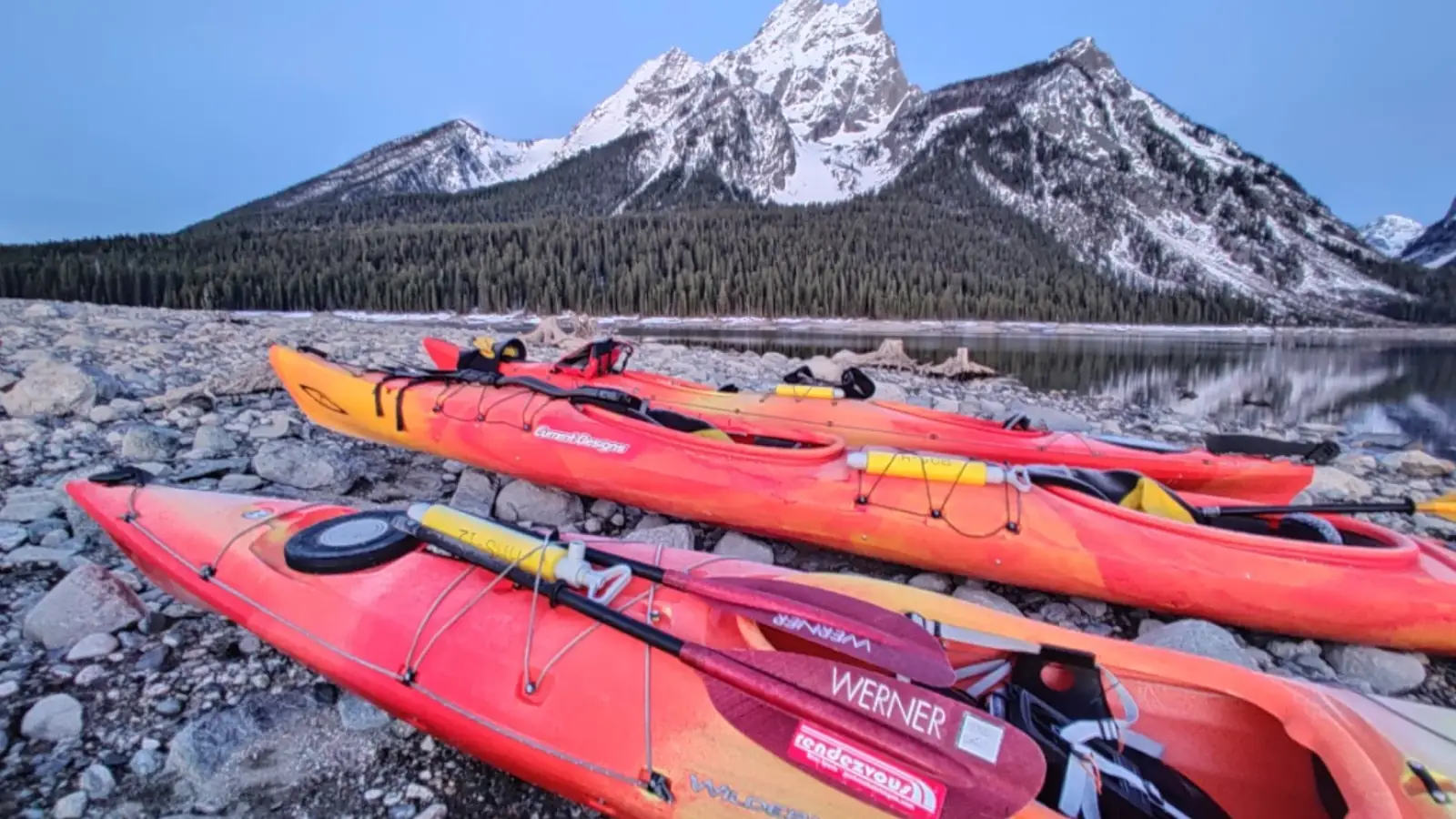 Multiple kayaks arranged on rocky shoreline and at least 8 kayaks in red and orange colors in Grand Teton National Park.
