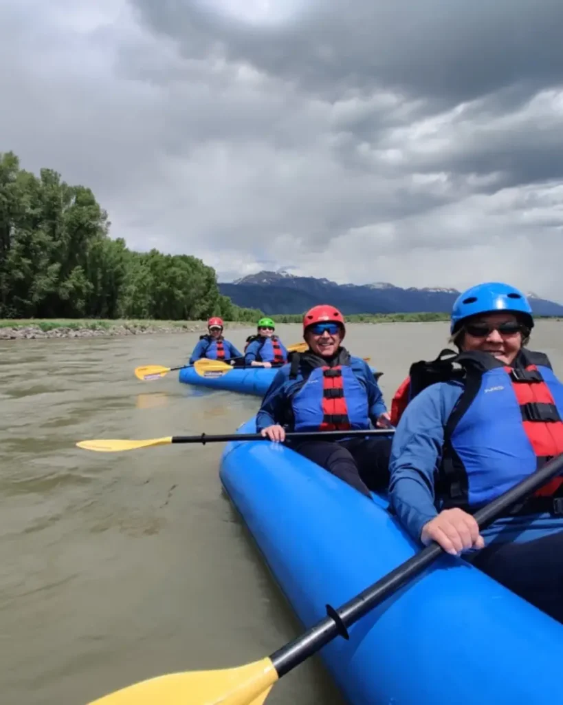 Five people in blue inflatable rafts and two rafts visible - foreground raft with two people, background raft with three people in Jackson Hole
