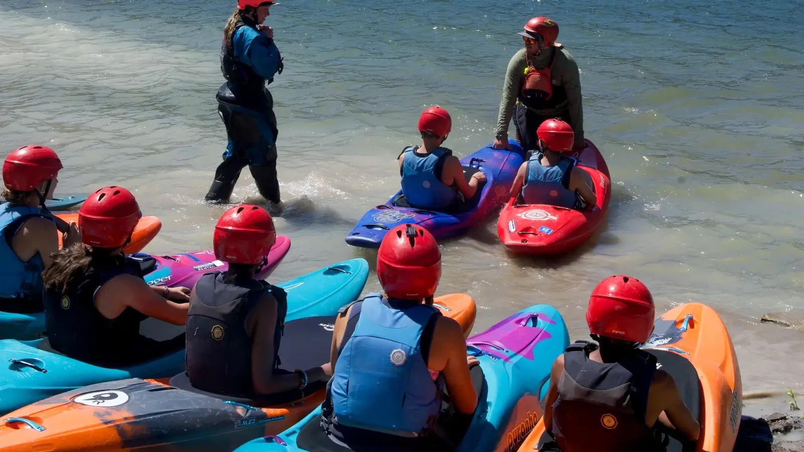 Group of people wearing red helmets and blue or black life vests seated in colorful kayaks at the shoreline and one adult standing in shallow water wearing a red jacket