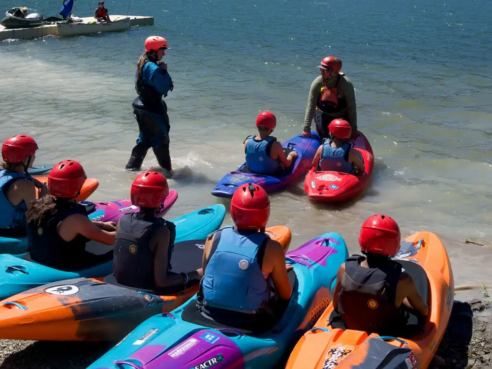 Group of people wearing red helmets and blue or black life vests seated in colorful kayaks at the shoreline and one adult standing in shallow water wearing a red jacket