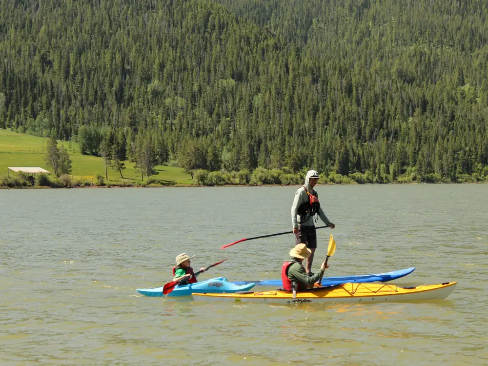 Three people on two kayaks on calm lake water and adult standing on blue paddleboard or kayak in background in Jackson Hole
