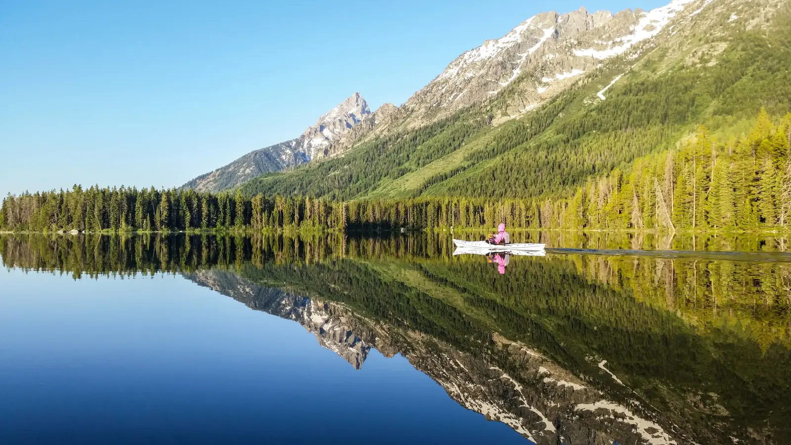 Calm lake with mirror-like reflection and mountain range with rocky peaks and patches of snow in Jackson Hole.