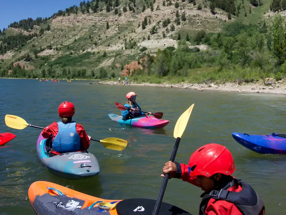 Three people in kayaks on a river and kayaks in orange or black, blue or gray, and pink or blue in Yellowstone National Park