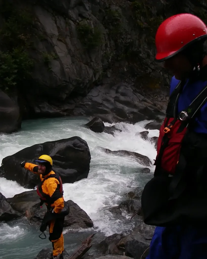 Two people on rocky riverbank and one person in foreground wearing a red helmet, blue outerwear, and black life vest in Jackson Hole