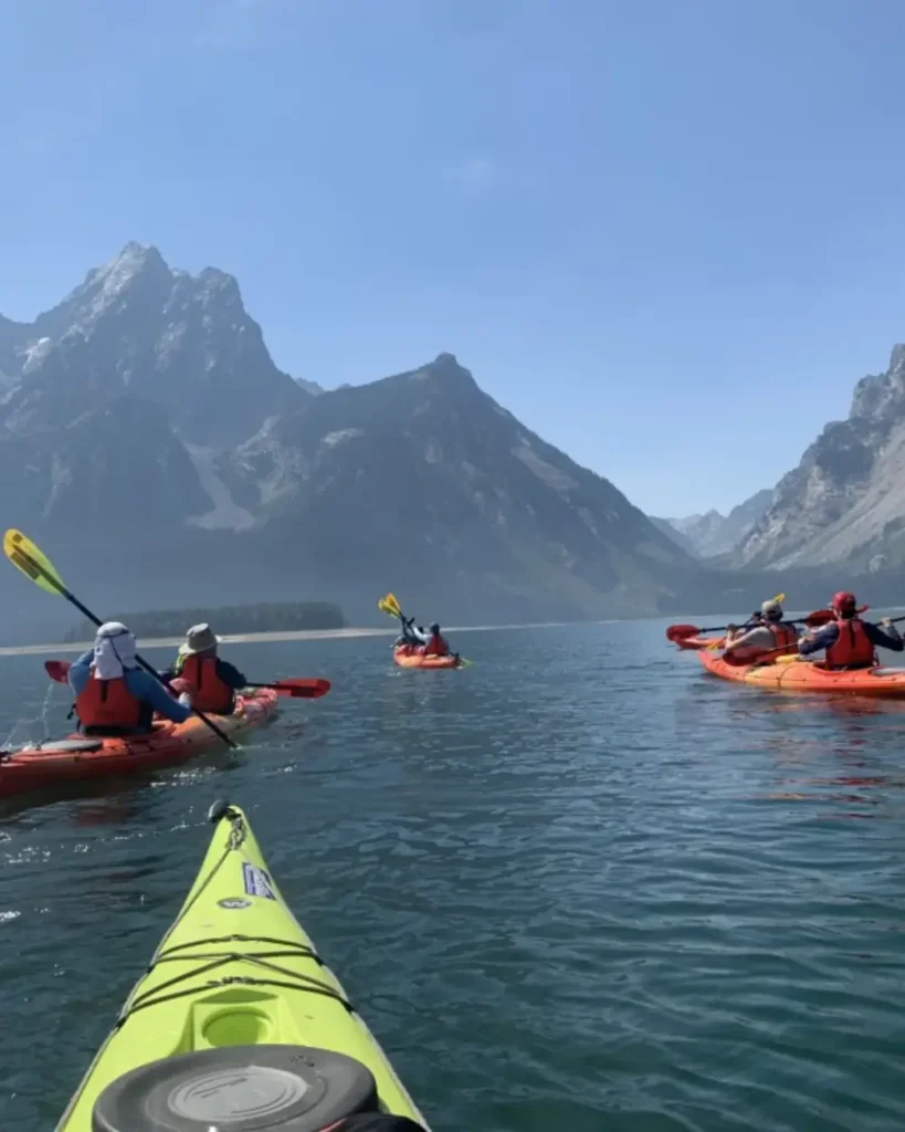Bow of a green kayak in the foreground with bungee cords and a round hatch and three orange kayaks on the water ahead in Snake River