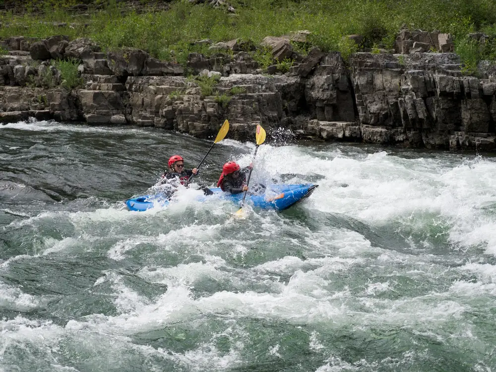 Two people in a blue inflatable kayak and both people wearing red helmets in Snake River.