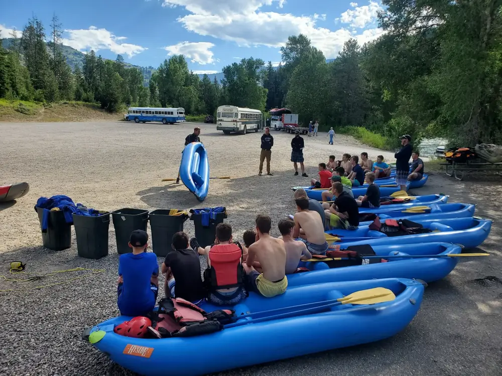 Multiple blue inflatable kayaks on gravel shore and group of people sitting in kayaks in Snake River.