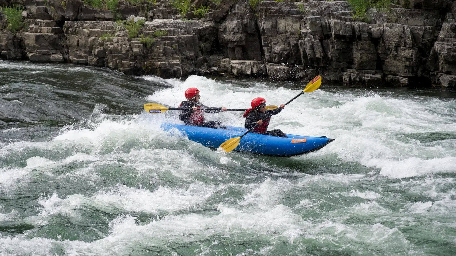 Two people in a blue inflatable raft and both people wearing red helmets and dark outerwear in Snake River.
