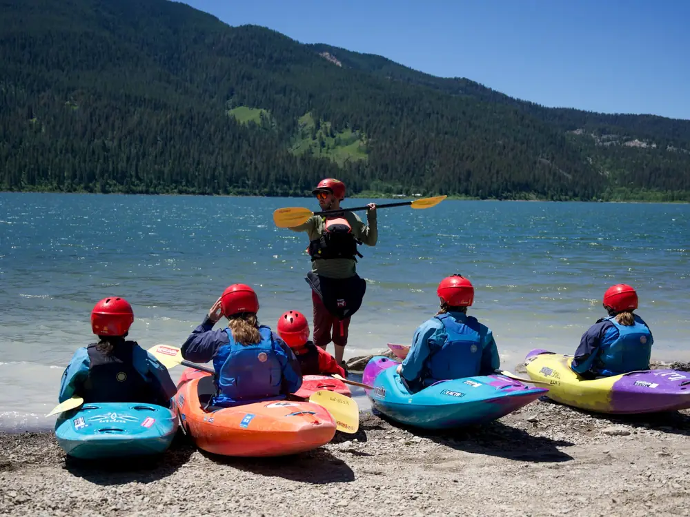 Six people wearing red helmets and life jackets and five kayaks on a rocky shoreline in blue, orange, purple or blue, and yellow colors in Jackson Hole