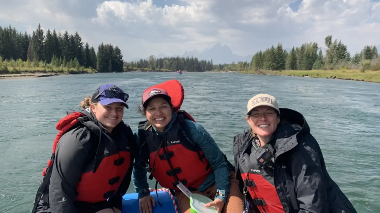 Three people on inflatable raft and all wearing PFDs in red and gray colors in Snake River.