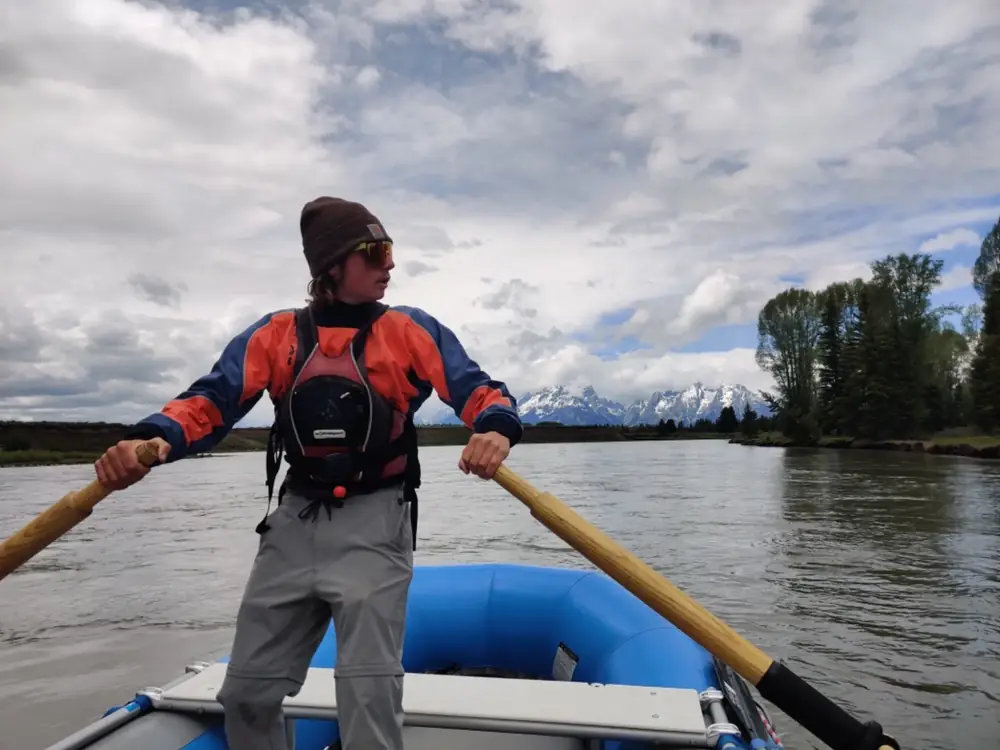 Person standing in blue inflatable raft and person wearing orange and blue dry top in Snake River.