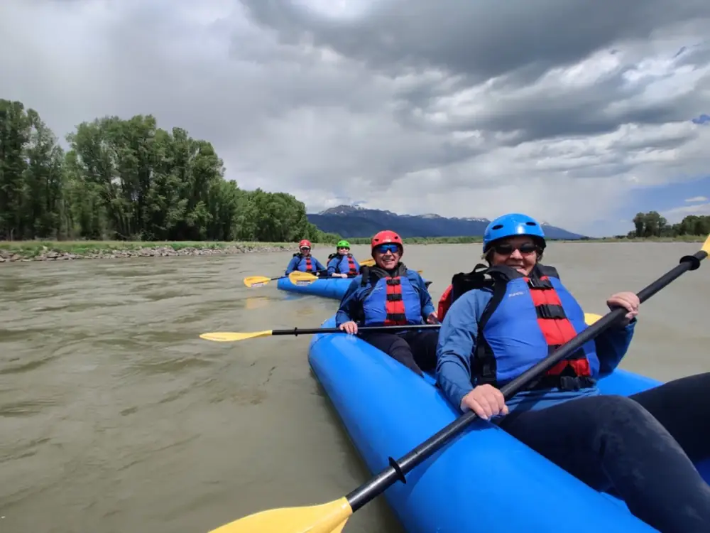 Five people in blue inflatable rafts and two rafts visible - foreground raft with two people, background raft with three people in Snake River