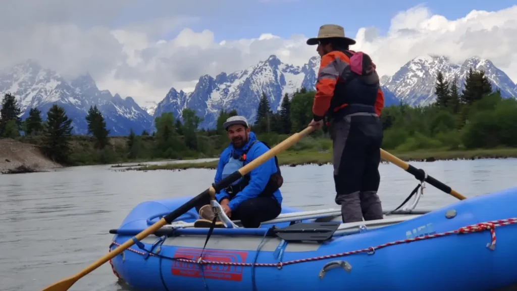 Two people in blue inflatable raft and person seated wearing blue shirt and black shorts in Jackson Hole.