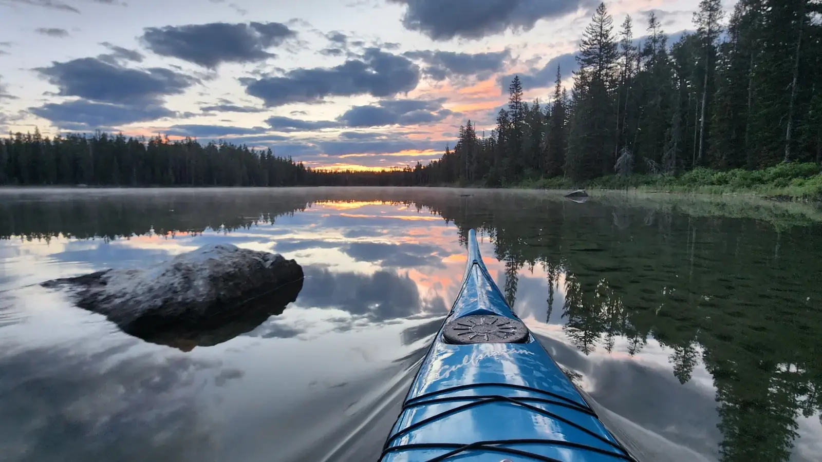 Blue kayak bow in the foreground with deck bungees and calm lake with mirror-like reflections of clouds and trees in Jackson Hole