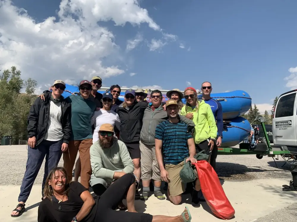 Group of approximately 16 people posing in parking lot and blue inflatable rafts on vehicle rack in background in Jackson Hole