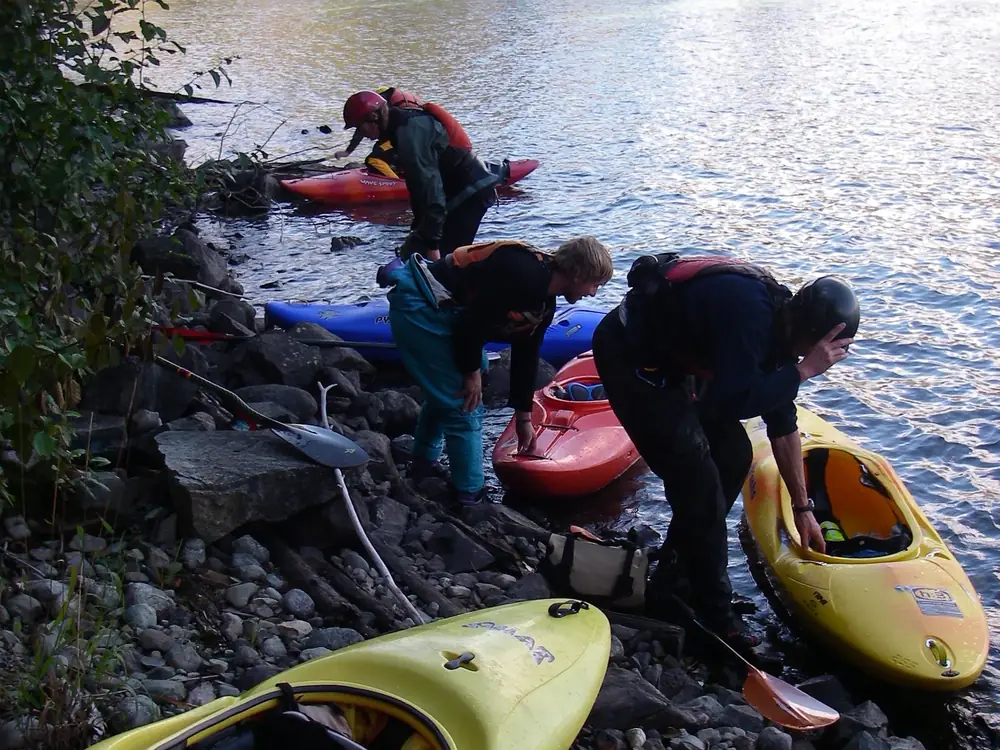 Three people on a rocky shoreline wearing paddling helmets and life vests and five kayaks on shore and in the water in Jackson Hole