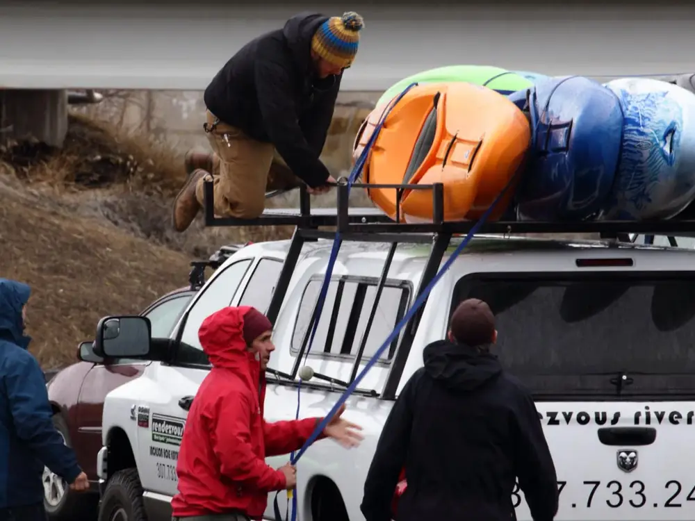 Four people near and on a white pickup truck and one person kneeling on a roof rack in Jackson Hole.