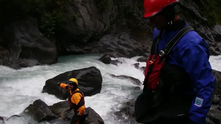 Two people on rocky riverbank and one person in foreground wearing a red helmet, blue outerwear, and black life vest in Jackson Hole.