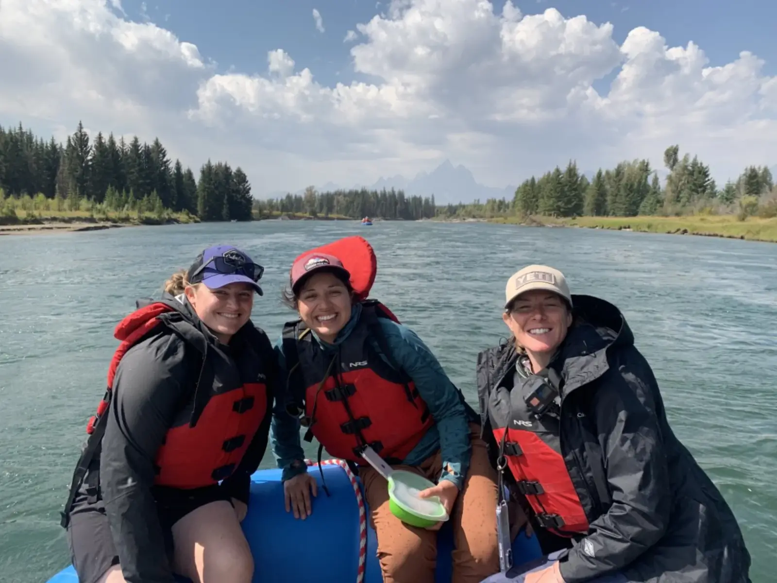 Three people on inflatable raft and all wearing PFDs in red and gray colors in Snake River.