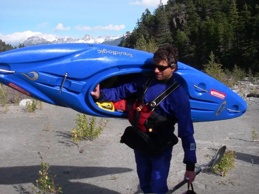 One person carrying a blue kayak on their right shoulder and person wearing a blue dry suit or waterproof suit with a red vest and black harness or strap in Jackson Hole