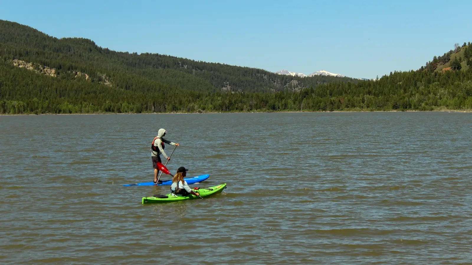 Two people on a body of water and one person standing on a blue paddleboard using a paddle with a red blade in Jackson Hole