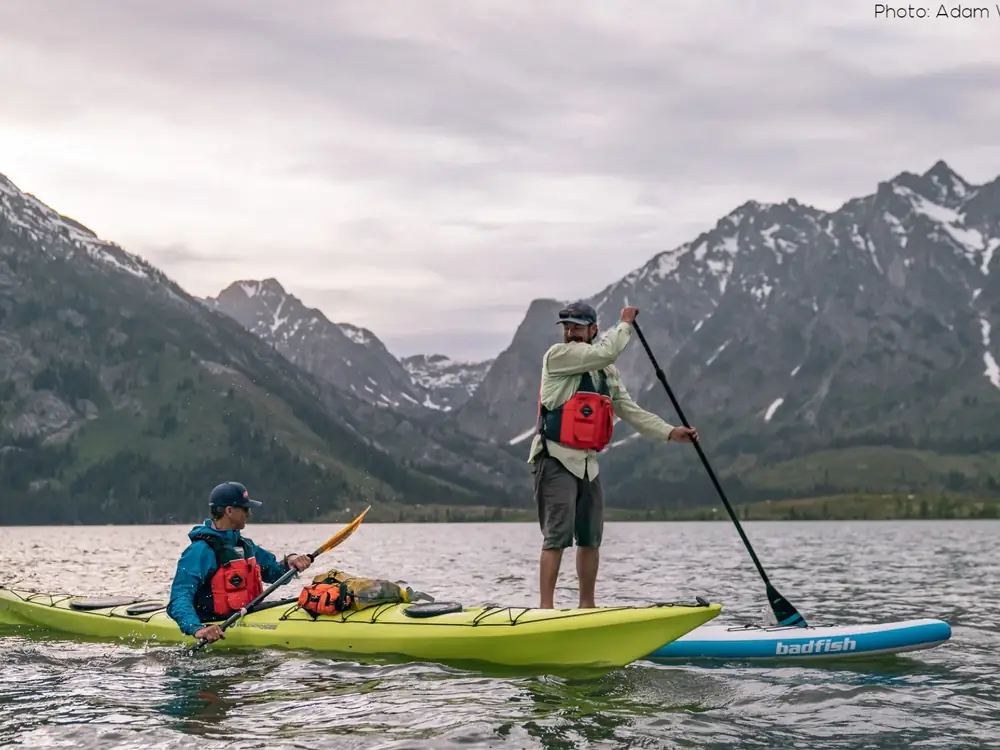 Person paddle boarding on calm water and red kayak in foreground in Jackson Hole.