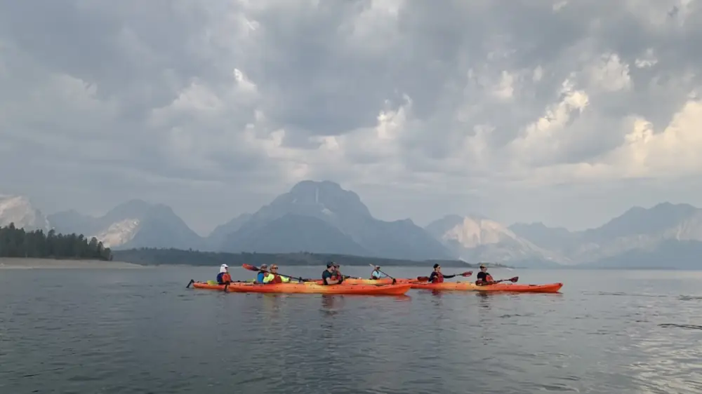Six people in orange kayaks and calm lake water in Yellowstone National Park.