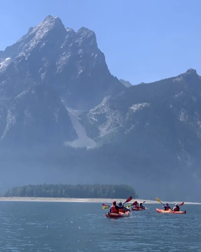 Three kayaks on calm lake water and red tandem kayak with two paddlers in center in Yellowstone National Park.