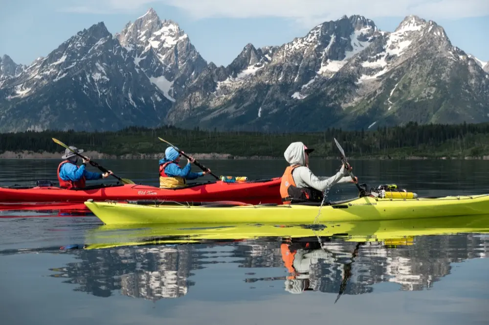 Three kayakers paddling on glassy calm Jackson Lake with Teton Range reflected in the water.